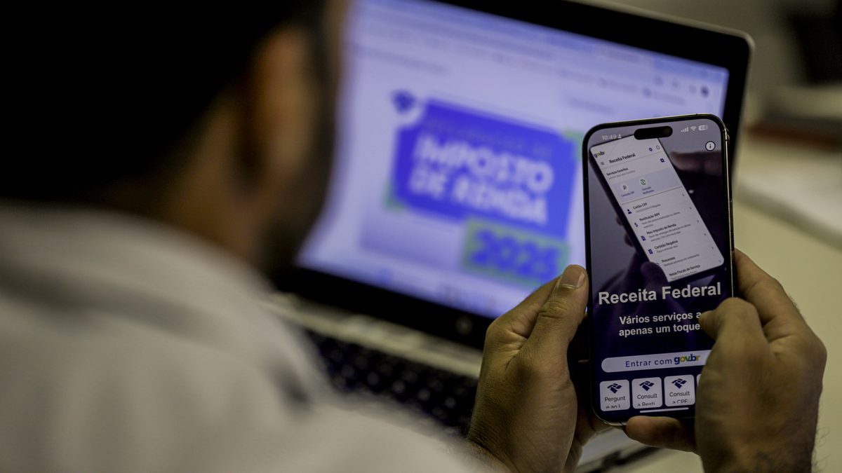 Contribuinte abre o aplicativo da Receita Federal no celular, com a página do Imposto de Renda no computador ao fundo