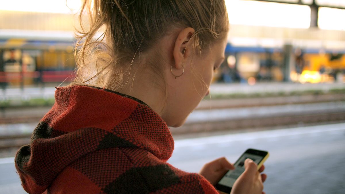 Mulher de costas, de blusa xadrez vermelha, olha o celular na plataforma de uma estação de trem