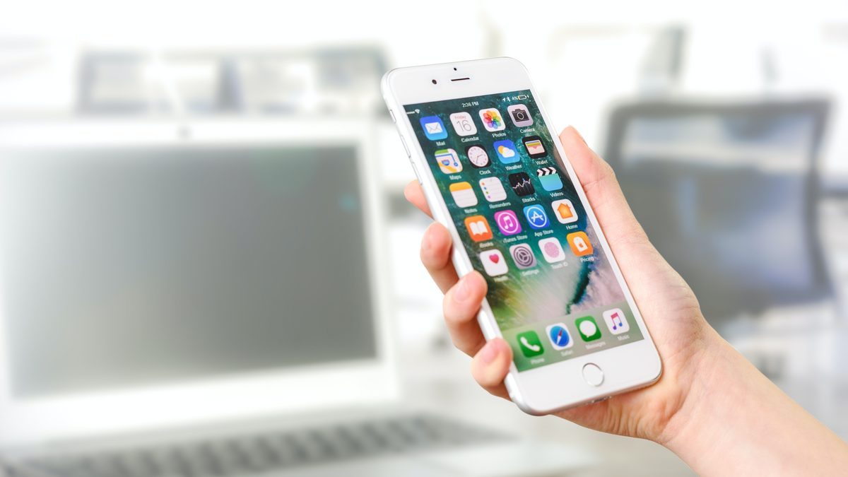 Mão segura um celular branco com a tela inicial cheia de ícones de apps, com um notebook desfocado ao fundo