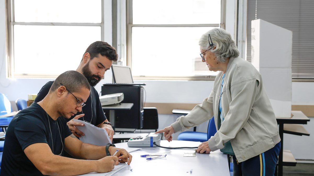 Eleitora idosa em frente à mesa de mesários, com a urna eletrônica ao lado, em seção eleitoral na UERJ, campus Maracanã, no Rio de Janeiro, no domingo da votação