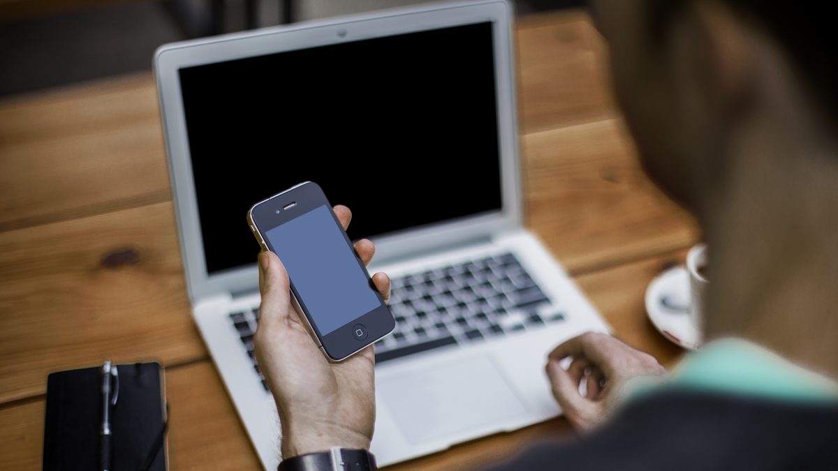 Pessoa com celular na mão diante de um notebook aberto