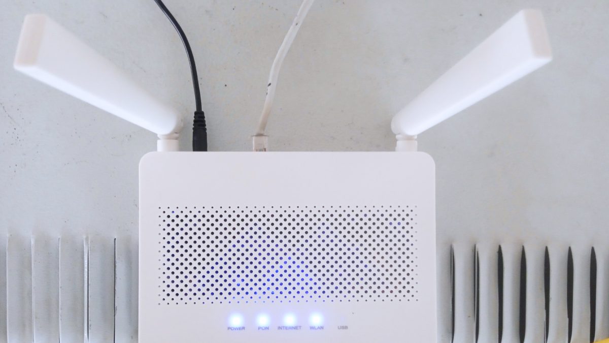 Roteador Wi-Fi branco com duas antenas e luzes azuis acesas, ligado a cabos sobre uma mesa clara